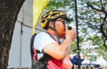 Petugas Jakarta Running Festival 2026 mengenakan helm sepeda dan rompi merah muda sambil menggunakan radio komunikasi Hytera untuk mendukung koordinasi selama ajang lari.
