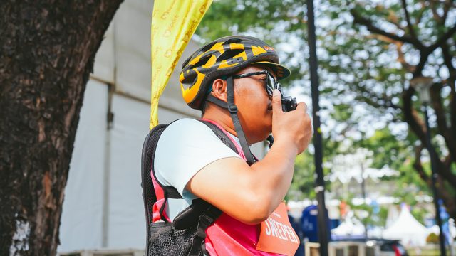 Petugas Jakarta Running Festival 2026 mengenakan helm sepeda dan rompi merah muda sambil menggunakan radio komunikasi Hytera untuk mendukung koordinasi selama ajang lari.