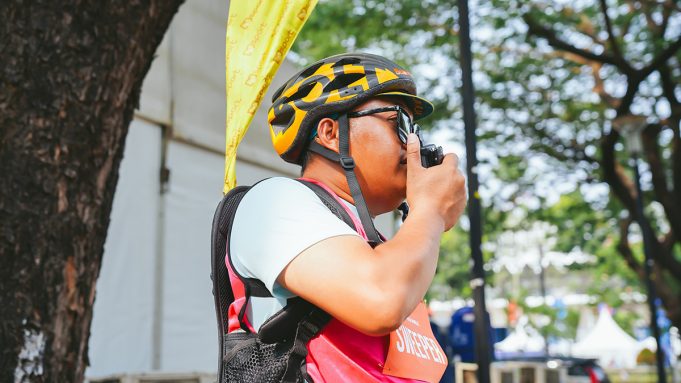 Petugas Jakarta Running Festival 2026 mengenakan helm sepeda dan rompi merah muda sambil menggunakan radio komunikasi Hytera untuk mendukung koordinasi selama ajang lari.